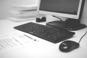 Eine schwarze Computertastatur und -maus auf einem weißen Schreibtisch, ein Stift liegt auf gedruckten Dokumenten vor einem Monitor, auf dem ein geöffnetes Dokument mit dem Titel „Anstiftung zum Mord“ zu sehen ist. Im Hintergrund ist ein Stapel Papiere zu sehen, wodurch das insgesamt schwarz-weiße Thema beibehalten wird.