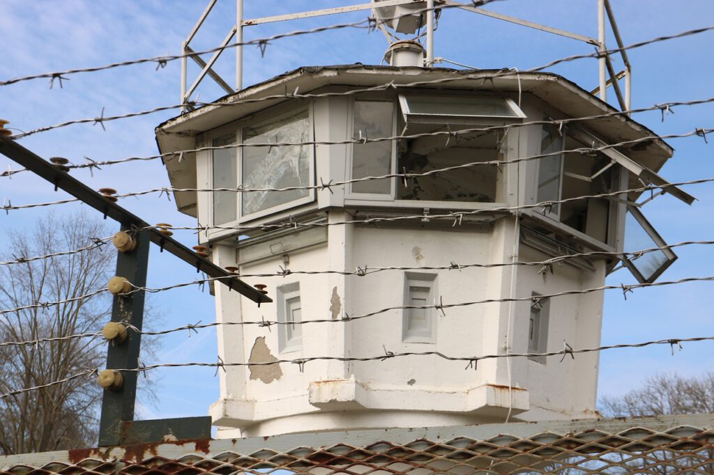 Hinter einem Stacheldrahtzaun steht unter einem klaren blauen Himmel ein weißer Wachturm mit zerbrochenen Fenstern. Der Turm weist Anzeichen von Verfall auf, die Farbe blättert ab. Im Vordergrund sind eine zweite Zaunschicht und freiliegende Kabel zu sehen. Im Hintergrund sind Bäume zu sehen.