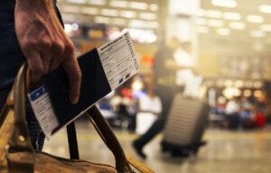 Eine Nahaufnahme einer Person, die in einer Hand eine Bordkarte und einen Reisepass hält und in der anderen eine Handtasche trägt. Im Hintergrund ist ein geschäftiges Flughafenterminal mit einer verschwommenen Gestalt zu sehen, die einen Koffer hinter sich herzieht.