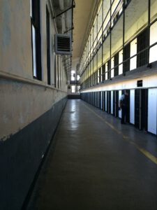 Ein langer, leerer Gefängniskorridor mit zwei Zellenreihen auf beiden Seiten. Das Bild zeigt eine einzelne Gestalt, die neben einer der Zellen auf der rechten Seite steht. Der Korridor ist schwach beleuchtet, natürliches Licht fällt durch die Fenster auf der linken Seite.