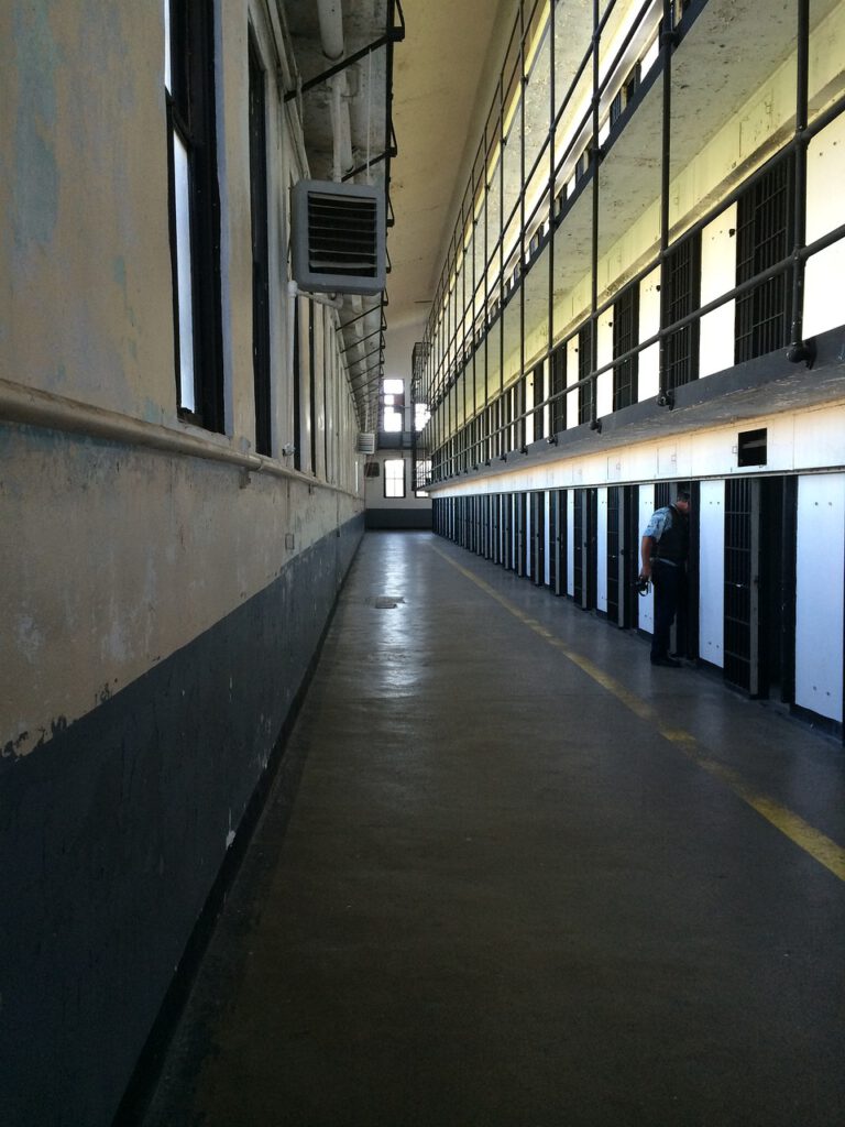 Ein langer, leerer Gefängniskorridor mit zwei Zellenreihen auf beiden Seiten. Das Bild zeigt eine einzelne Gestalt, die neben einer der Zellen auf der rechten Seite steht. Der Korridor ist schwach beleuchtet, natürliches Licht fällt durch die Fenster auf der linken Seite.