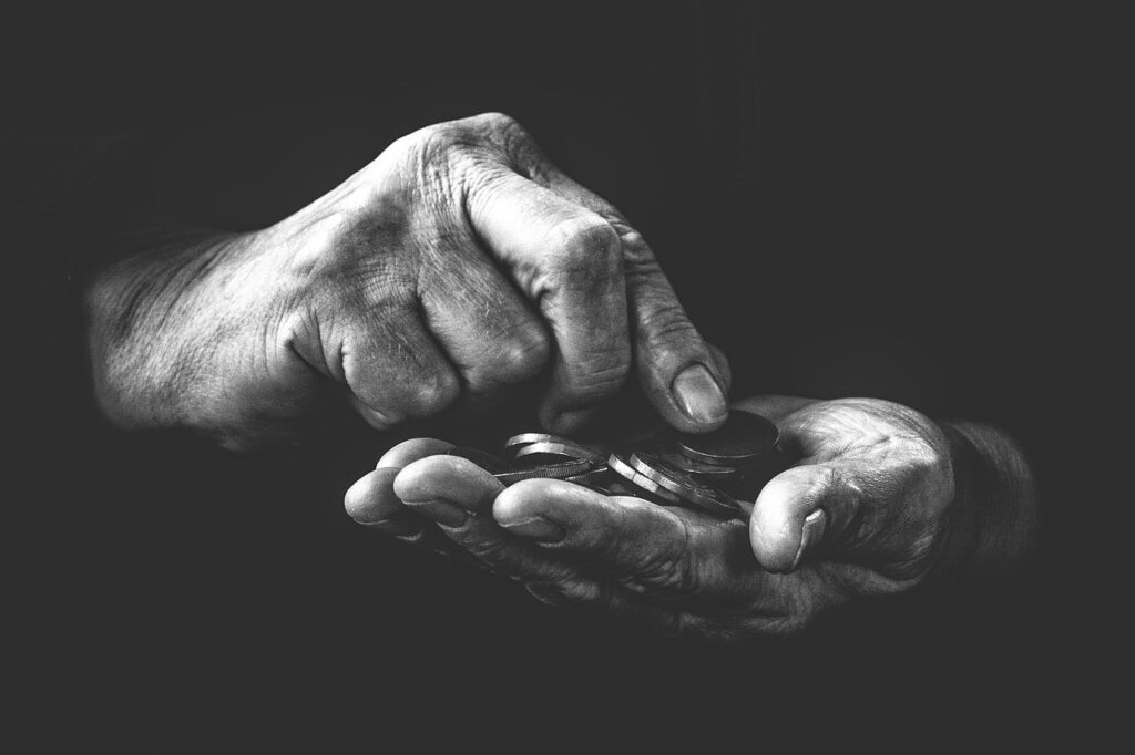 Nahaufnahme in Schwarzweiß von Händen, die einen kleinen Stapel Münzen halten und zählen. Eine Hand ist offen und die Münzen liegen in der Handfläche, während die Finger der anderen Hand die Münzen durchsehen. Der Hintergrund ist dunkel.