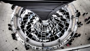 Blick von oben in eine moderne Architekturstruktur mit geschwungenen Linien und reflektierenden Oberflächen. Menschen gehen und betrachten ausgestellte Fotos. Das Bild erzeugt aufgrund der Reflexionen und Kurven eine symmetrische, fast schwindelerregende Perspektive.