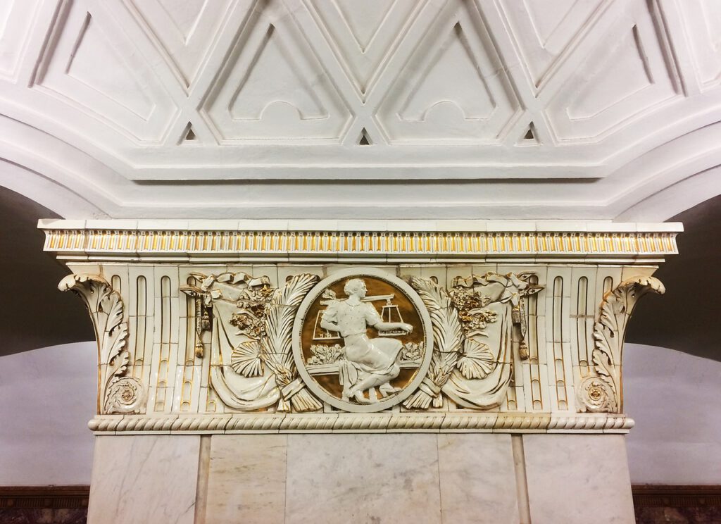 Ein kunstvolles architektonisches Detail mit einer Flachreliefskulptur einer Person, die eine Waage hält, ist in ein kompliziertes, klassisches Design eingebettet, das florale und geometrische Elemente enthält. Die Decke darüber ist mit symmetrischen, rautenförmigen Mustern geschmückt.