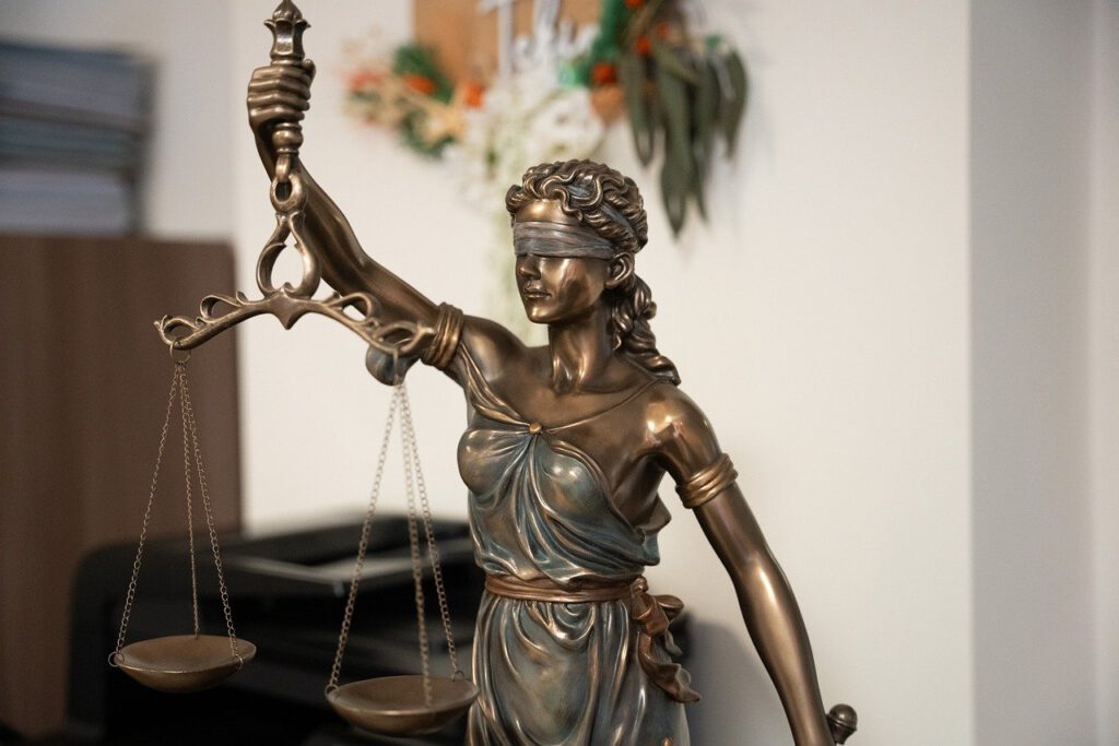 Ein Bild einer Bronzestatue der Justitia. Die Statue zeigt eine Frau mit verbundenen Augen, die in ihrer linken Hand eine Waage und in ihrer rechten Hand ein Schwert hält. Im Hintergrund ist eine verschwommene Bürokulisse mit einigen Papieren und Dekorationen zu sehen.