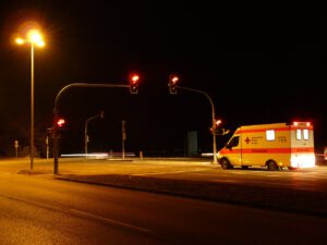 Ein gelber Krankenwagen steht nachts an einer roten Ampel an einer Kreuzung. Die Straße wird von hohen Straßenlaternen beleuchtet und der Hintergrund ist dunkel. Die Straße ist größtenteils leer und die Lichter der fahrenden Fahrzeuge sind als Streifen sichtbar.