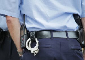 Ein uniformierter Polizist ist von hinten zu sehen. Der Polizist hat ein Funkgerät, Handschellen und eine Waffe im Holster am Gürtel. Die blaue Uniform besteht aus einem kurzärmeligen Hemd und einer dunklen Hose.
