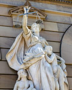 Eine Steinstatue stellt Justitia dar. In ihrer linken Hand hält sie eine Waage und mit ihrem rechten Arm den Rücken eines jungen Menschen. Eine sitzende Figur blickt von unten zu Justitia auf. Die Statue ist an einem Gebäude mit Steinmauern befestigt.