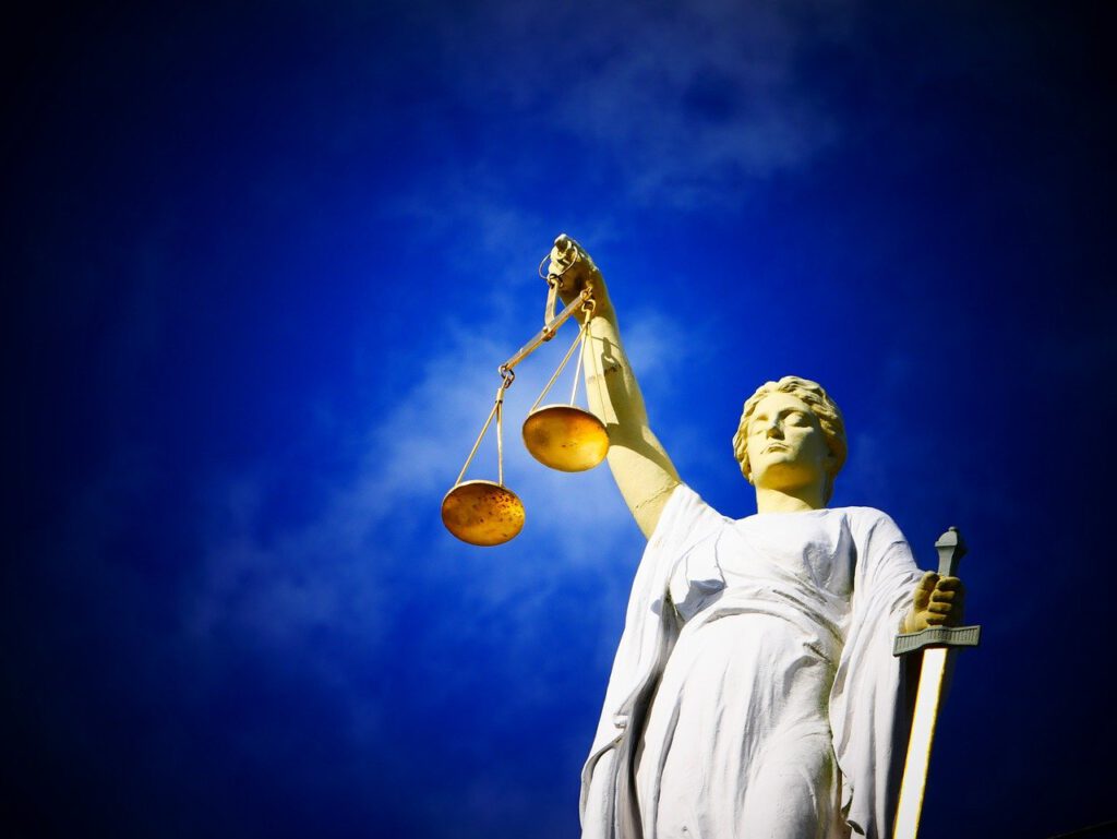 Vor einem strahlend blauen Himmel ist eine Statue der Justitia zu sehen. In ihrer linken Hand hält sie die Waage der Gerechtigkeit und in ihrer rechten ein Schwert. Sie trägt ein wallendes Gewand und blickt leicht nach oben.