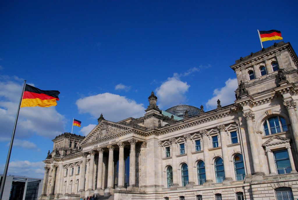 Das Bild zeigt das Reichstagsgebäude in Berlin, Deutschland, unter einem klaren blauen Himmel mit einigen Wolken. Deutsche Flaggen sind auf dem Gebäude und an Fahnenmasten im Vordergrund zu sehen. Die Architektur zeichnet sich durch klassische Säulen und detailliertes Mauerwerk aus.