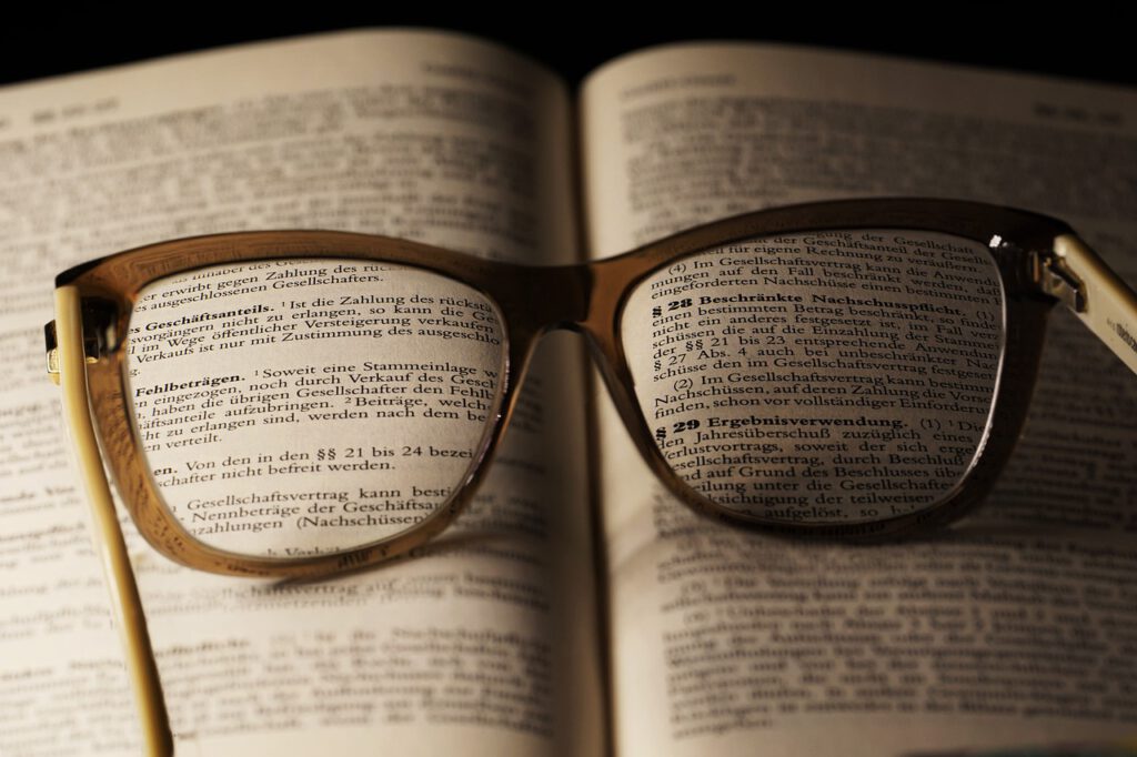 Eine Brille mit hellem Gestell liegt aufgeklappt auf einem Buch und vergrößert den Text auf den Seiten darunter. Die Gläser der Brille zeigen die gedruckten Wörter deutlich an und erleichtern so das Lesen. Im Mittelpunkt steht ein deutscher Text, der sich mit Der Fall Till Lindemann beschäftigt.