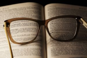 Eine Brille mit hellem Gestell liegt aufgeklappt auf einem Buch und vergrößert den Text auf den Seiten darunter. Die Gläser der Brille zeigen die gedruckten Wörter deutlich an und erleichtern so das Lesen. Im Mittelpunkt steht ein deutscher Text, der sich mit Der Fall Till Lindemann beschäftigt.