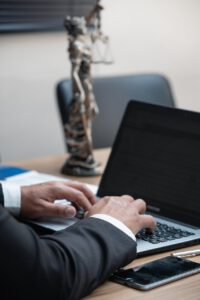 Eine Person im Anzug tippt an einem Schreibtisch auf einem Laptop. Auf dem Schreibtisch liegen außerdem ein Smartphone und eine Justitia-Statue. Der Hintergrund ist unscharf.