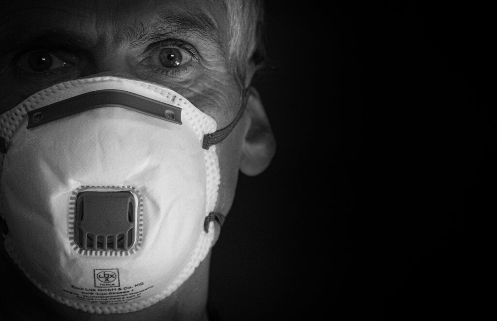Ein Graustufenbild einer Person, die eine Schutzmaske mit Ventil trägt, die Nase und Mund bedeckt. Der Hintergrund ist dunkel und nur die Hälfte des Gesichts der Person ist sichtbar, wodurch ihr Auge und die Textur der Maske hervorgehoben werden.