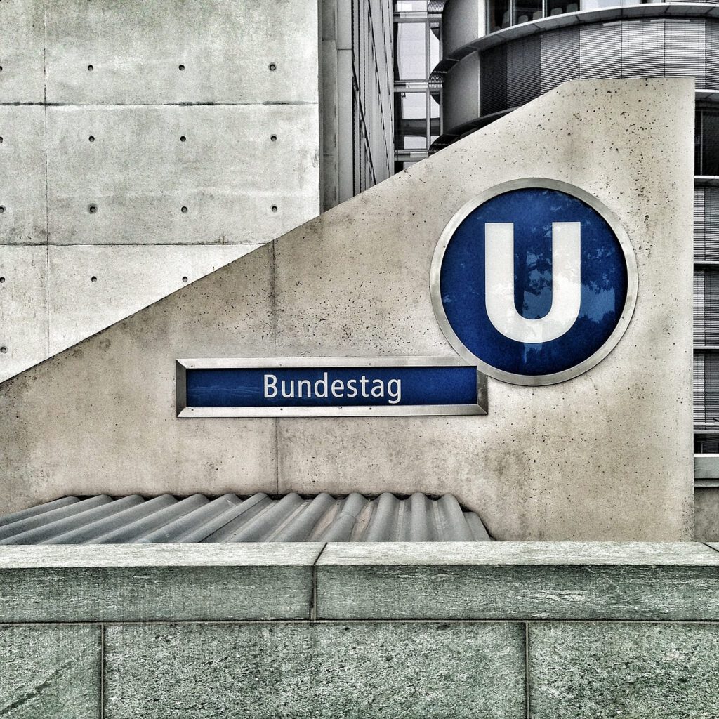 An einer Betonwand ist ein blau-weißes U-Bahn-Schild mit dem Buchstaben „U“ und dem Wort „Bundestag“ zu sehen, das den Eingang zu einer U-Bahnstation markiert. Im Hintergrund sind moderne Bürogebäude zu sehen.
