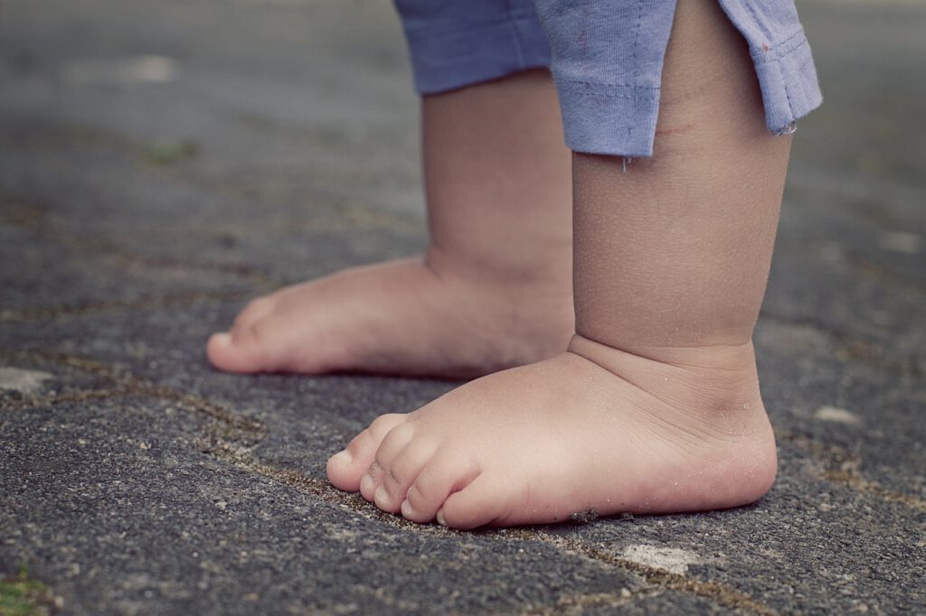 Eine Nahaufnahme der nackten Füße eines Babys, das auf einer gepflasterten Fläche steht. Das Baby trägt hellblaue Hosen, die leicht hochgekrempelt sind, und die Füße liegen nebeneinander. Der Belag besteht aus Ziegeln in verschiedenen Schattierungen.