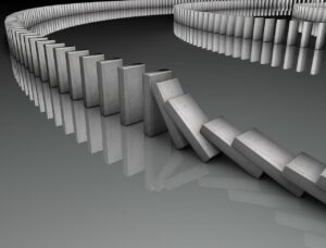 Eine Reihe grauer Dominosteine ist in einer gekrümmten Linie auf einer reflektierenden Oberfläche angeordnet. Mehrere Dominosteine haben begonnen umzufallen, wodurch ein Dominoeffekt entsteht. Der Hintergrund ist ein Farbverlauf von Dunkel- zu Hellgrau.
