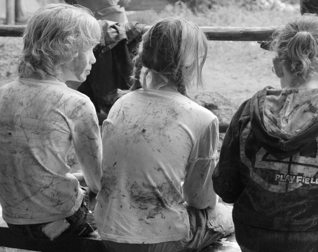 Unfälle auf Klassenfahrten -Drei Kinder sitzen mit dem Rücken zur Kamera auf einer Bank. Sie tragen schmutzige Kleidung und scheinen draußen gespielt zu haben. Die Umgebung wirkt natürlich und rustikal, mit Holzelementen um sie herum.
