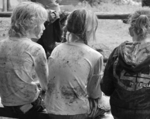Unfälle auf Klassenfahrten -Drei Kinder sitzen mit dem Rücken zur Kamera auf einer Bank. Sie tragen schmutzige Kleidung und scheinen draußen gespielt zu haben. Die Umgebung wirkt natürlich und rustikal, mit Holzelementen um sie herum.