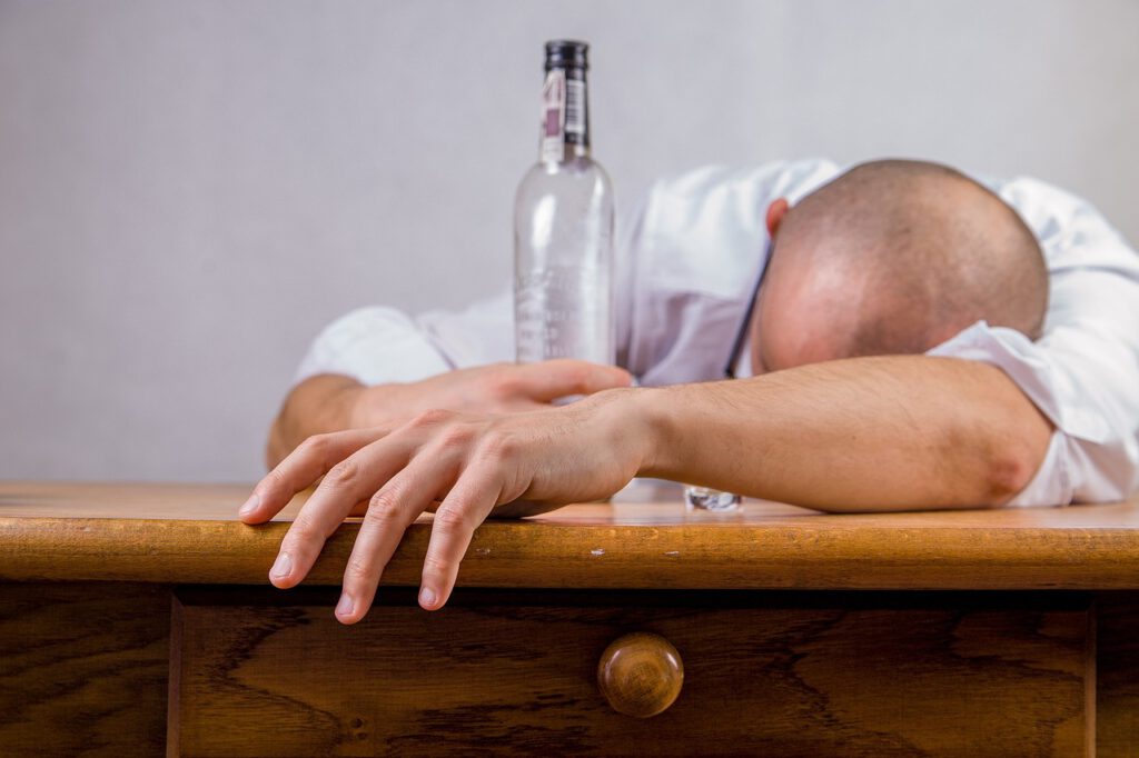 Eine Person mit rasiertem Kopf sitzt zusammengesunken über einem Holztisch und hält in der ausgestreckten Hand eine durchsichtige Flasche. Sie scheint ein weißes Hemd zu tragen.