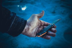 Die Hand einer Person hält ein Klappmesser mit einem schwarz-orangen Griff. Der Hintergrund ist eine verschwommene, blau getönte Oberfläche, möglicherweise Schnee oder Eis.