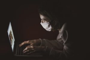 Eine Person mit Maske sitzt in einem schwach beleuchteten Raum und benutzt einen Laptop. Das Licht des Bildschirms beleuchtet ihr Gesicht, während sie sich intensiv auf den Kampf gegen Online-Verleumdung konzentriert. Ihre Hände ruhen auf der Tastatur, bereit, schädliche Falschinformationen abzuwehren.