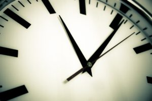 Nahaufnahme eines Zifferblatts. Ein zeitlicher Bezugspunkt zeigt die Uhrzeit ungefähr mit 10:08 Uhr an. Die Uhr hat schwarze Stunden- und Minutenzeiger, einen braunen Sekundenzeiger und schwarze Stundenmarkierungen auf makellos weißem Hintergrund.