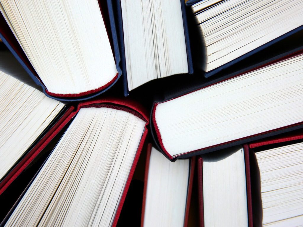 Eine Nahaufnahme mehrerer Bücher, die mit dem Rücken nach außen angeordnet sind und so ein kreisförmiges Muster bilden. Die Seiten sind aufgefächert und geben den Blick auf das weiße Papier und einige rote Akzente auf den Buchdeckeln frei.