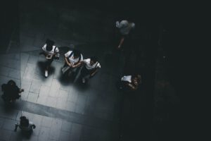 Eine Luftaufnahme zeigt fünf Menschen, die auf einem grauen, gefliesten Bürgersteig gehen. Die meisten von ihnen sind weiß und blau gekleidet. Eine Person ist in ihr Telefon vertieft. Der schwach beleuchtete Bereich, der an die Feierlichkeit der Jugendrichterlichen Weisungen erinnert, wirft Schatten auf die Oberfläche.