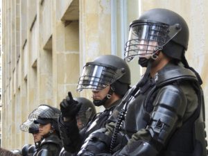 Vier Personen in Kampfausrüstung, Helme und Gesichtsschutz, stehen wachsam vor einem Gebäude. Sie gehören wahrscheinlich einem Sicherheitsdienst oder der Polizei an und sind einsatzbereit, während die Diskussionen über die Polizeikosten bei Fußballspielen weiterhin großes Interesse und Debatten auslösen.