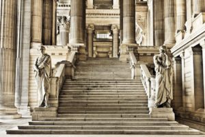 Eine große Treppe mit verziertem Steingeländer führt zwischen großen klassischen Säulen nach oben und symbolisiert die komplexe Struktur des Strafverfahrens. Zwei steinerne Statuen in Roben stehen am Fuße der Treppe und vermitteln ein Gefühl von Würde und den komplexen Ausländerrechten, die dieser imposanten Architektur zugrunde liegen.