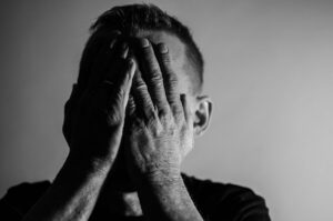 Ein Schwarz-Weiß-Bild einer Person, die ihr Gesicht mit den Händen bedeckt. Sie trägt kurze Haare und ein dunkles Hemd. Der Hintergrund ist schlicht und weist einen Farbverlauf von Hell nach Dunkel auf.